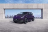 Three-quarter front view of a purple Nissan PIXO electric city car parked beneath a modern concrete structure, with a city skyline in the background.